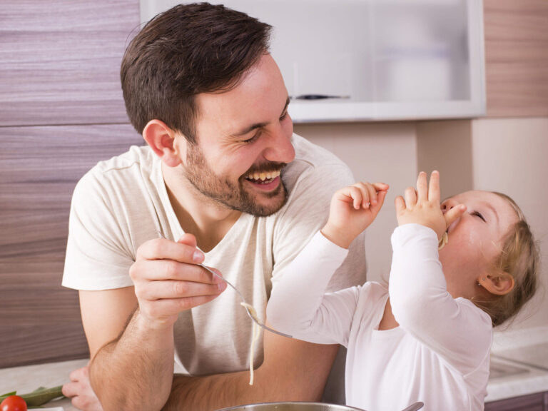 Homem branco de cabelos pretos curtos e barba curta, vestindo camiseta branca, pai de uma bebê branca e de cabelos claros, também de camiseta branca. ela coloca o macarrão na boca com a cabeça virada para trás enquanto o pai sorri e segura o garfo.